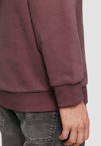 Bordeauxrotes Sweatshirt mit geripptem Bündchen und Saum, aus weichem Stoff, auf einem grauen Hintergrund präsentiert, mit einer Hand, die an der Seite ruht.