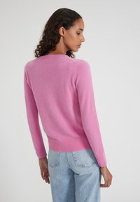 Donna con capelli ricci che indossa un maglione rosa a maniche lunghe e jeans azzurri chiari, di profilo contro uno sfondo bianco semplice.