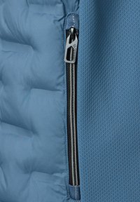 Blauer isolierter Jackenstoff mit einem gesteppten Muster links und glatter Textur rechts. Verfügt über einen silbernen Reißverschluss mit schwarzem Zuglasche.