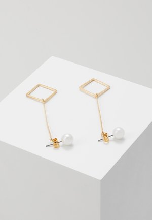 Gouden vierkante draad oorbellen met dunne kettingen die enkele witte parel stekers vasthouden op een witte geometrische ondergrond.