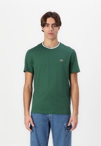Tricou verde din bumbac, cu guler rotund, având un tiv alb contrastant, mâneci scurte și un mic logo brodat pe piept.