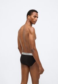 Μαύρο σλιπ με γκρι ελαστική ζώνη που φέρει το λογότυπο "TOMMY HILFIGER", εμφανιζόμενο από το πλάι σε γυμνό κορμό.