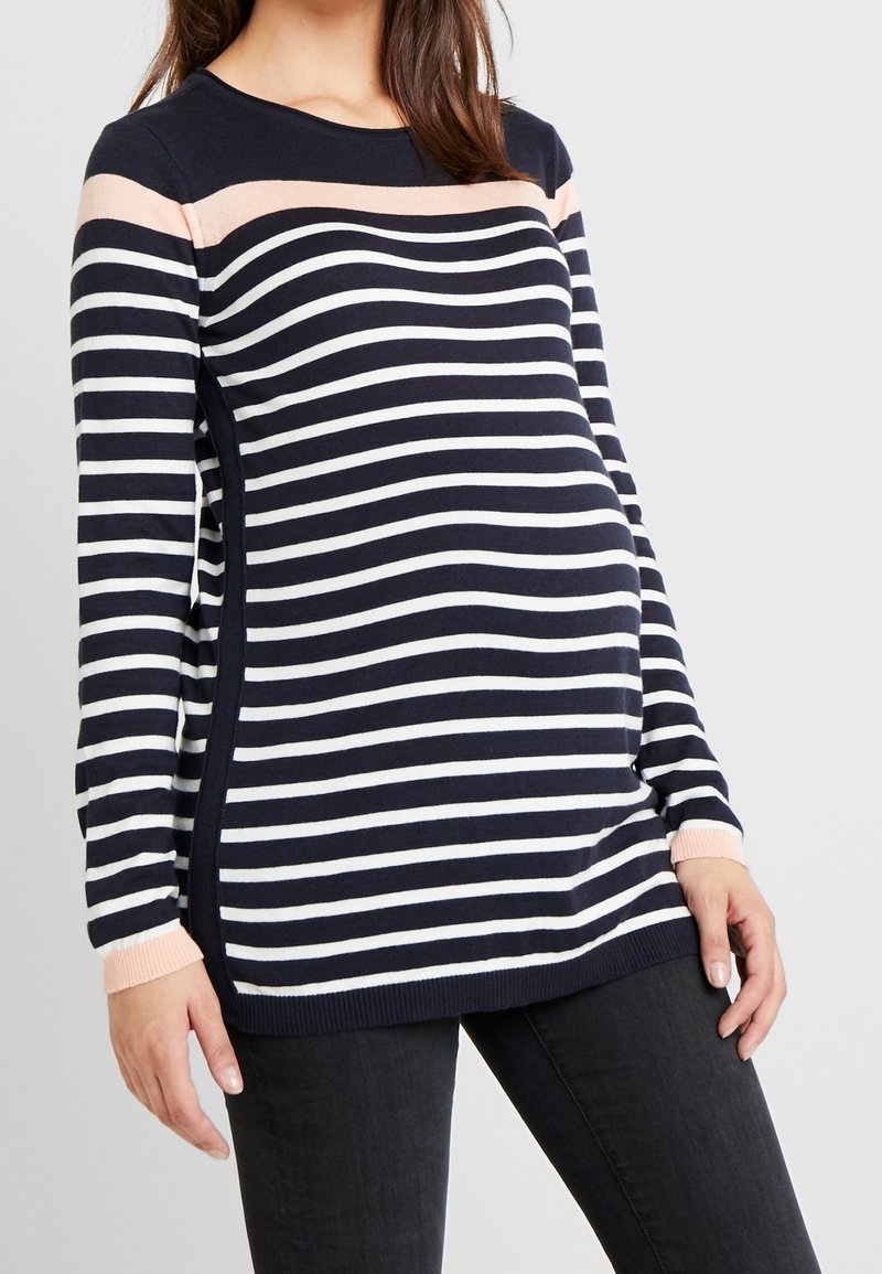 Femme enceinte portant un pull à rayures horizontales bleu marine et blanc avec des accents rose clair, associé à un pantalon noir, debout devant un fond gris.
