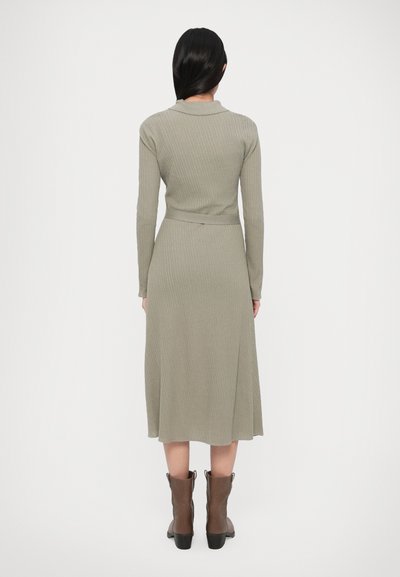 Robe côtelée à manches longues de couleur vert olive, cintrée à la taille avec une ceinture. L'ourlet arrive au niveau des genoux, portée avec des bottines marron.