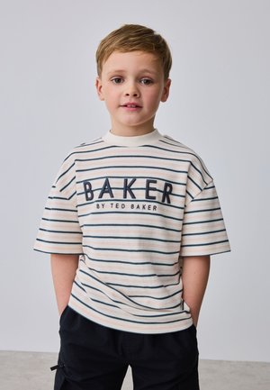 Noor poiss lühikeste blondide juustega, kandes triibulist T-särki tekstiga "BAKER BY TED BAKER" ja musti pükse, seisab tavalise halli tausta ees.