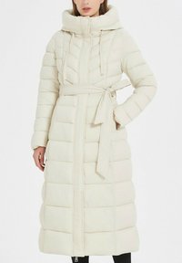Manteau matelassé beige clair avec une taille cintrée, une fermeture éclair sur toute la longueur, une grande capuche et des coutures en chevrons rembourrées. Texture douce et manches longues.