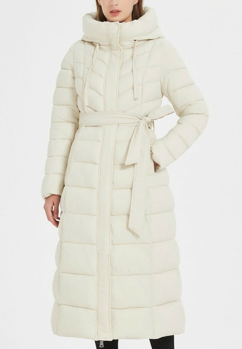 Manteau matelassé beige clair avec une taille cintrée, une fermeture éclair sur toute la longueur, une grande capuche et des coutures en chevrons rembourrées. Texture douce et manches longues.