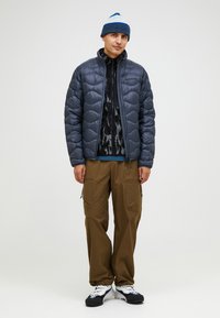 Grau gesteppte Jacke mit Wellenmuster, schwarze Reißverschluss-Schicht, braune Cargo-Hosen und blauer Beanie. Die Schuhe sind schwarz mit weißen Akzenten.