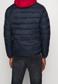 Mand iført en marineblå kviltet jakke med rød hætte og mørkegrå jeans, står med ryggen til mod en ensfarvet baggrund.