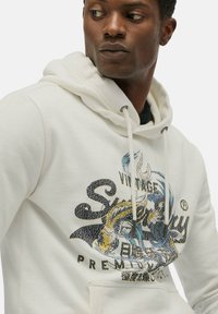 Sudadera blanca de tela suave con un bolsillo frontal, que presenta un estampado gráfico vintage colorido y un detalle de cordón en la capucha.