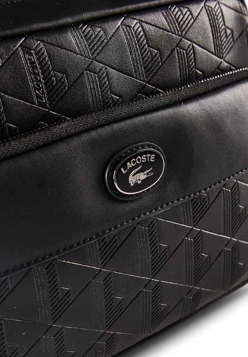 Black textured leather bag with embossed geometric patterns and a metal oval Lacoste logo featuring a crocodile.