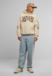 Krem hettegenser med grafisk logo, sammen med lyseblå baggy jeans og hvite sneakers. Kappen har et kamuflasjemønster med en logo.