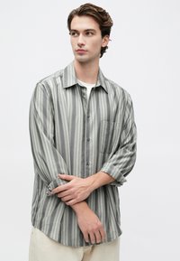 Joven con cabello castaño ondulado que lleva una camiseta de manga larga a rayas grises sobre una camiseta blanca, ajustándose las mangas mientras mira hacia un lado.