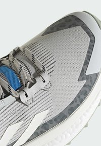 Jasnoszary but sportowy z teksturowaną siateczką na cholewce, niebieskim akcentem i wzorzystym szarym nakładką. Posiada białe paski ozdobne oraz gumową podeszwę.