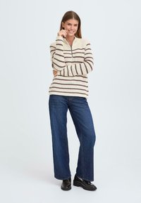 Gestreifter Strickpullover in Creme mit dunkelbraunen horizontalen Streifen, ausgestattet mit einem Reißverschlusskragen und langen Ärmeln, kombiniert mit weit geschnittenen Denim-Hosen.