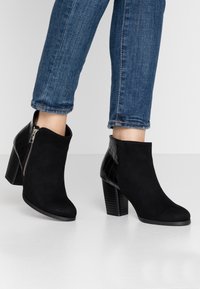 Sorte ankelstøvler med overdel i ruskind, krokodillepræget hæl, lynlås i siden og en stablet hældesign. Parret med opfoldede blå jeans.