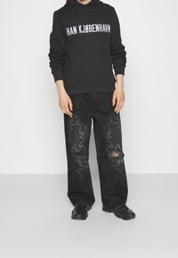 Felpa nera con cappuccio e la scritta "HAN KJØBENHAVN", abbinata a jeans neri con un motivo testurizzato e dettagli consumati. Sneakers nere.