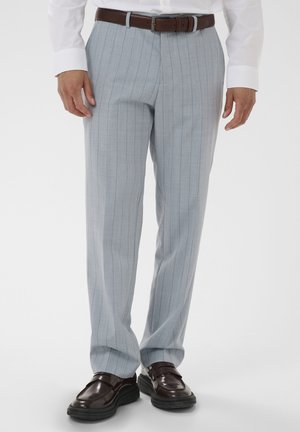 Mand iført lysegrå bukser med nålestriber, mørkebrunt bælte, hvid skjorte og mørkebrune lædermokkasiner, stående foran hvid baggrund.