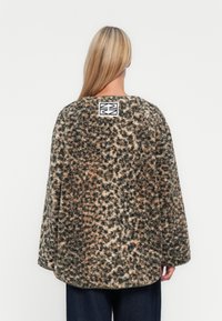 Ξανθιά γυναίκα φοράει ένα χαλαρό, φuzzy σακάκι με λεοπάρ σχέδιο και με μια μαύρη και λευκή μπάλα στην άνω πλάτη, στέκεται μπροστά σε απλό φόντο.