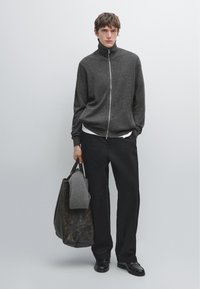 Grå zip-up tröja med hög krage, svarta vida byxor och en stor mörk väska innehållande en hopvikt grå stickad tröja. Slät textur.
