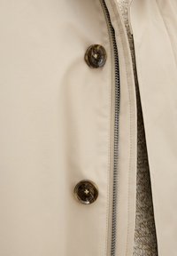 Giacca beige con zip e due bottoni scuri, marmorizzati; texture liscia con un interno a maglia visibile.
