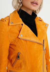 Oranje suede motorjacket met een notch-kraag, zilverkleurige hardware en ritszakken. Textuurafwerking voegt dimensie toe.