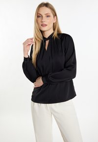 Schwarze Seidenbluse mit langen Ärmeln, Schlüsselloch-Ausschnitt und gerafften Details an Schultern und Bündchen, kombiniert mit cremefarbenen Hosen.