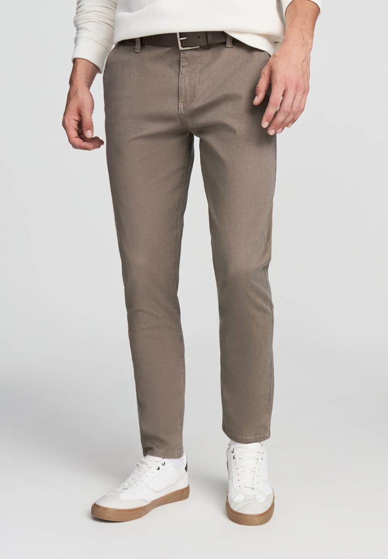 Chino slim-fit marroni con texture e finitura liscia, abbinati a sneakers bianche e una cintura scura, dotati di tasche anteriori e un look sartoriale.
