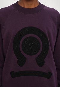 Fialová mikina s vyvýšeným čiernym logom s textom "SAVAGE FURY IN A THUNDERSTORM" a "VERSACE JEANS COUTURE," s raglanovými rukávmi.