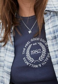 Chemise bleu marine à manches courtes avec logo brodé blanc et motif de laurier, portée sous une chemise en flanelle à carreaux claire. Collier en argent avec détail.