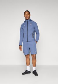 Tuta Nike blu composto da felpa con zip, tasche laterali e logo, abbinata a pantaloni corti coordinati e scarpe da ginnastica nere.