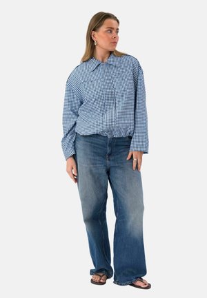 Femme debout, portant une chemise à carreaux bleue à manches longues, un jean bleu ample, des tongs noires et des boucles d'oreilles argentées, regardant sur le côté.