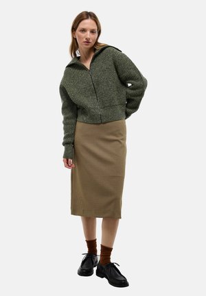 Veste zippée verte à texture avec une capuche, associée à une jupe marron à damier de longueur genoux. Le mannequin porte des chaussettes brunes et des chaussures noires.