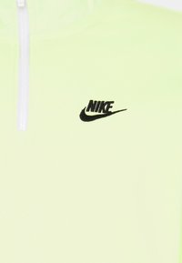 Nike-pullover i ljus neongrön, med en svart broderad logotyp på bröstet och en vit dragkedja. Slät textur och sportig design.