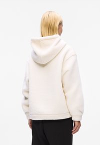 Valkoinen fleece-huppari rentoa istuvuutta, tilava huppu ja ribbireunat. Tekstuurissa on pehmeä tuntu, näkyvät ompeluyksityiskohdat.