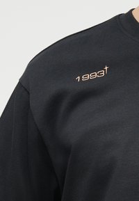 Μαύρο φούτερ με το «1993» τυπωμένο με μικρά μπεζ γράμματα κοντά στον αριστερό ώμο, με σχέδιο σταυρού ως μέρος του αριθμού.