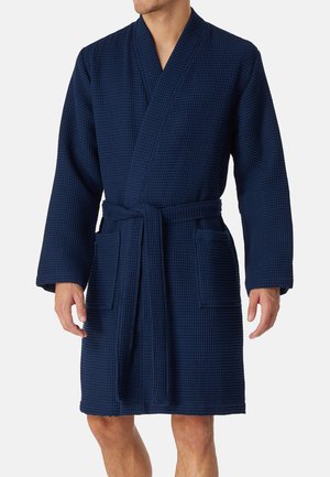 Mężczyzna ubrany w granatowy szlafrok o fakturze waflowej, sięgający do kolan, z długimi rękawami, dwoma przednimi kieszeniami i paskiem wiązanym w talii.