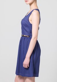 Hihaton, A-line mekko syvän sinisenä, jossa on tiukka yläosa ja kerrostettu hame, korostettuna ohuella metallivyöllä vyötäröllä.