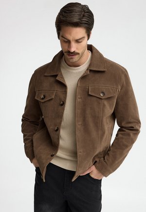 Hombre con una chaqueta de ante marrón sobre un suéter de canalé beige, con las manos en los bolsillos de los pantalones negros, mirando hacia abajo.