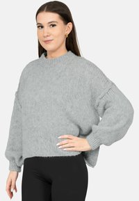 Jonge vrouw draagt een losse grijze gebreide trui en zwarte broek, staand met één hand op haar heup tegen een effen achtergrond.