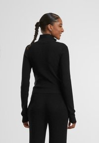 Sweater noir à col roulé côtelé avec des manches longues. Présente un design ajusté et un tricot texturé. La vue arrière montre un col montant et une silhouette épurée.