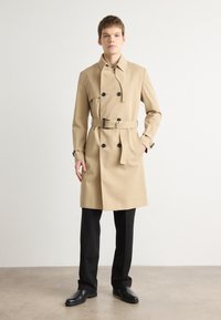 Caban bej cu un design cu două rânduri de nasturi, nasturi negri, curea ajustabilă și buzunare laterale, asociat cu pantaloni negri și pantofi.