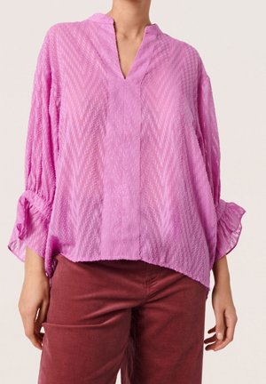 Kobieta ubrana w luźną, teksturowaną lawendową bluzkę z dekoltem w serek i rękawami 3/4, w połączeniu z wysokim stanem, rdzawe spodnie.