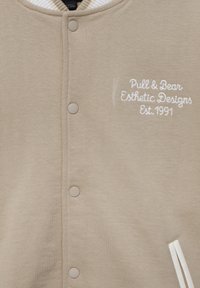 Veste beige à boutons avec col en côte blanc, texte brodé "Pull & Bear Esthetic Designs Est. 1991" et poche inclinée avec bordure blanche.