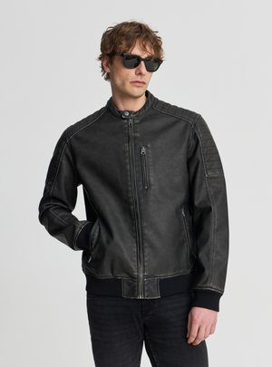 Uomo con giacca di pelle nera e occhiali da sole, in piedi con una mano in tasca davanti a uno sfondo neutro.