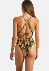 Maillot de bain une pièce noir avec un motif floral doré, présentant des bretelles croisées et un détail noué dans le dos. Texture douce, jambes à coupe haute.