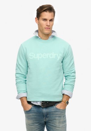 Sweatshirt bleu clair avec logo "Superdry", col et poignets côtelés, superposé sur une chemise rayée bleue, associé à un jeans en denim léger et une ceinture noire.