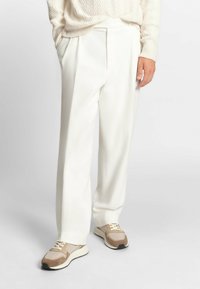 Pantaloni bianchi a gamba larga con una texture liscia e pieghe frontali, abbinati a sneakers beige e marroni con dettagli in rete e suede.