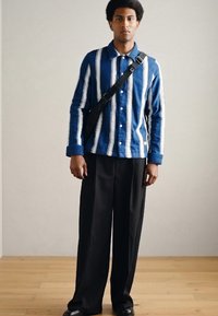 Niebiesko-biała koszula w paski na guziki z długimi rękawami, zestawiona z luźnymi czarnymi spodniami. Czarna torba na ramię oraz błyszczące czarne buty.