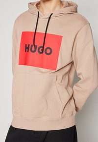 Sudadera con capucha beige que presenta un gráfico rectangular rojo llamativo con la palabra "HUGO" en negro. El material parece suave, y cuenta con un bolsillo frontal.